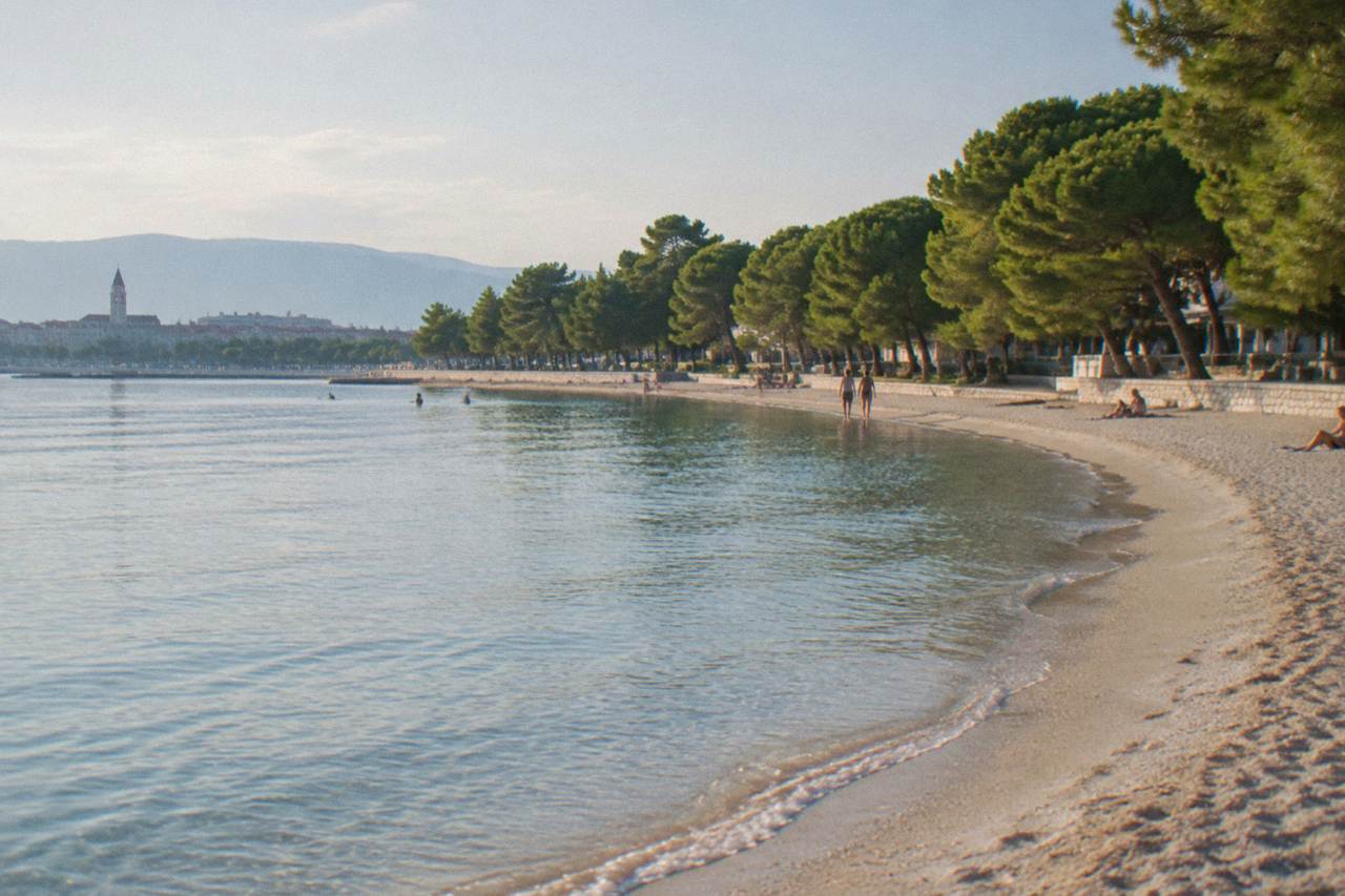 A sandy city beach in Split with calm shallow water and pine trees behind the shore