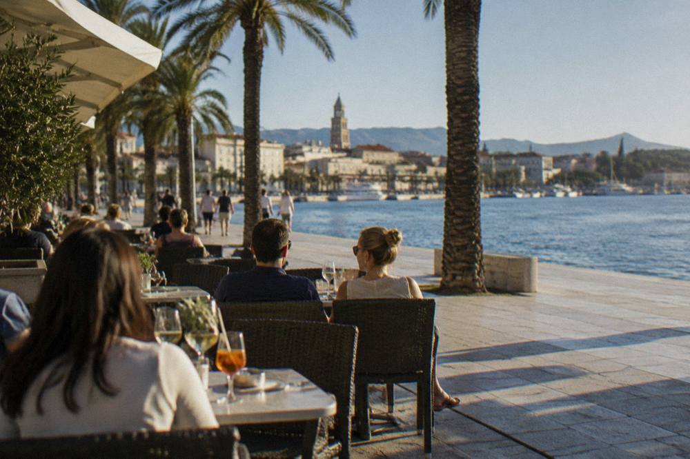 People seated with drinks at outdoor cafe tables along the seafront promenade in Split