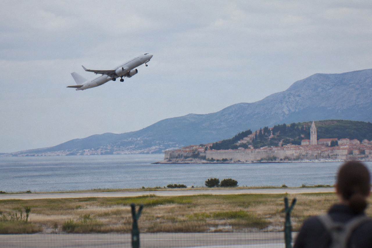 An airliner climbing above the Adriatic coastline near Split with wooded hills and a walled old town below