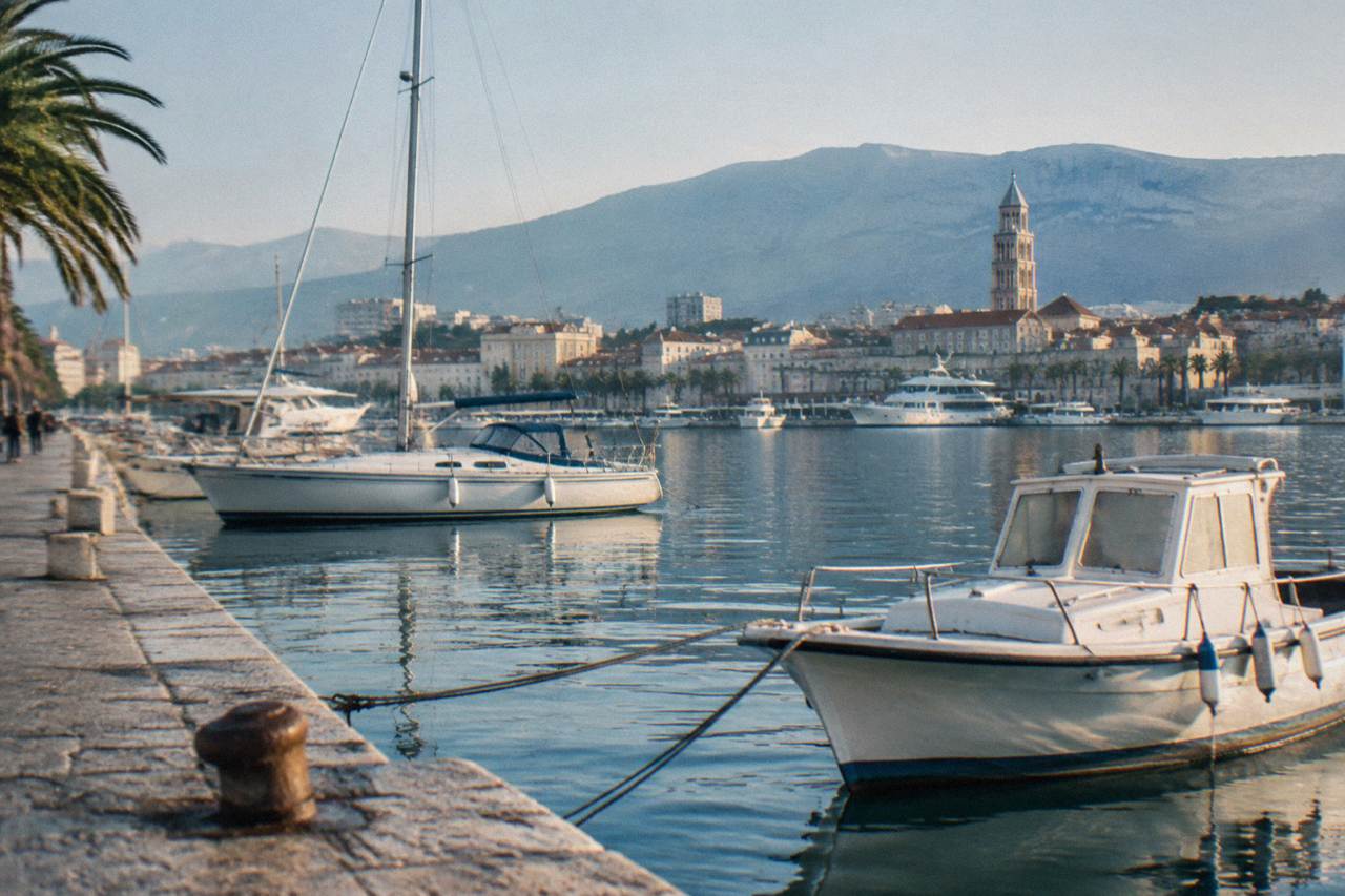 Boats moored in a stone harbour with hills rising behind a Dalmatian coastal town