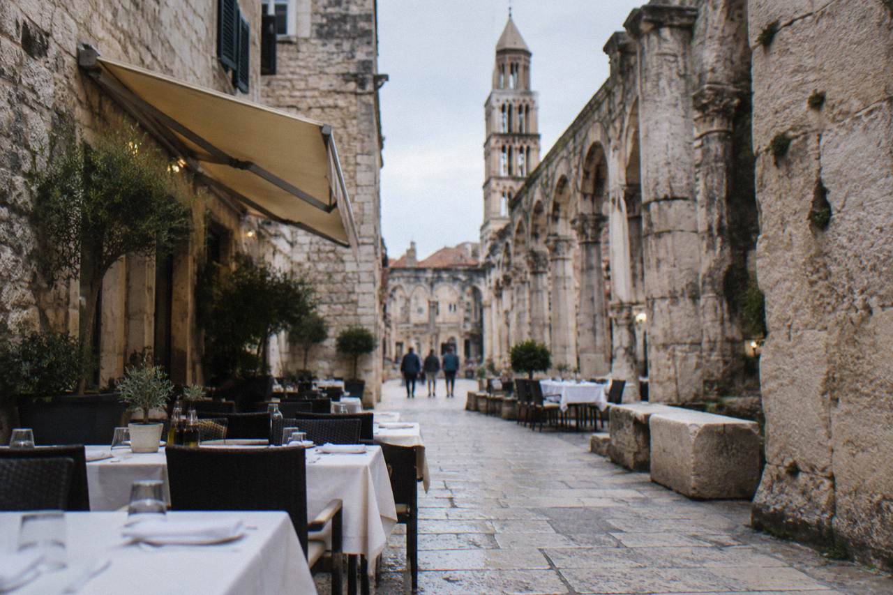 Outdoor restaurant tables with white tablecloths set along a stone street in a Mediterranean old town