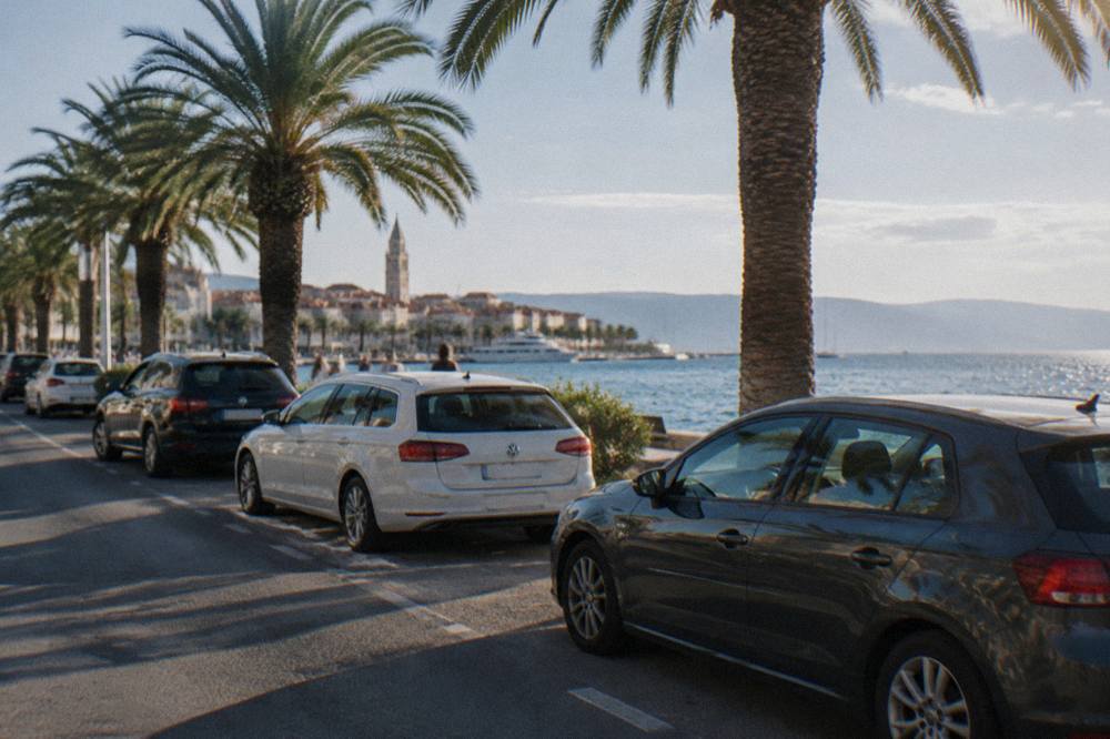 Parked cars along a coastal road with palm trees and the Adriatic Sea behind them