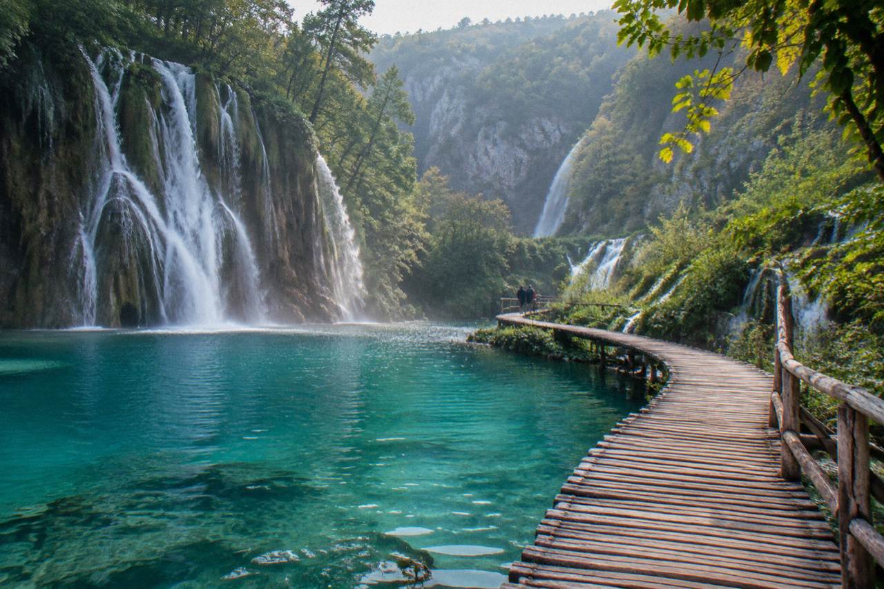 Wooden footbridge crossing a turquoise lake between waterfalls in a forested canyon