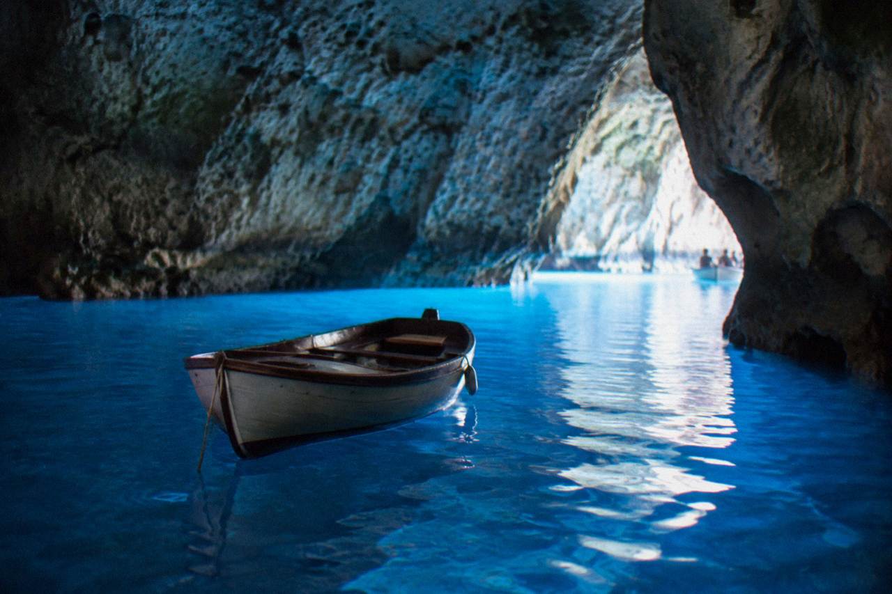 A small wooden boat floating inside a sea cave lit by blue light reflecting off the water