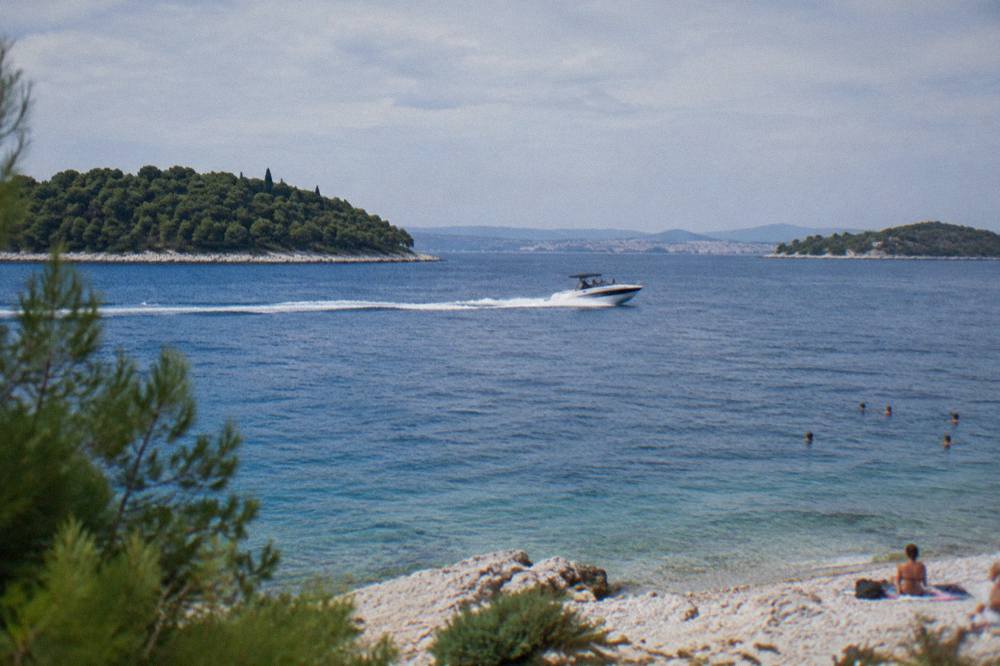 A speedboat crossing open blue water between wooded islands near Split with swimmers near a small pebble beach