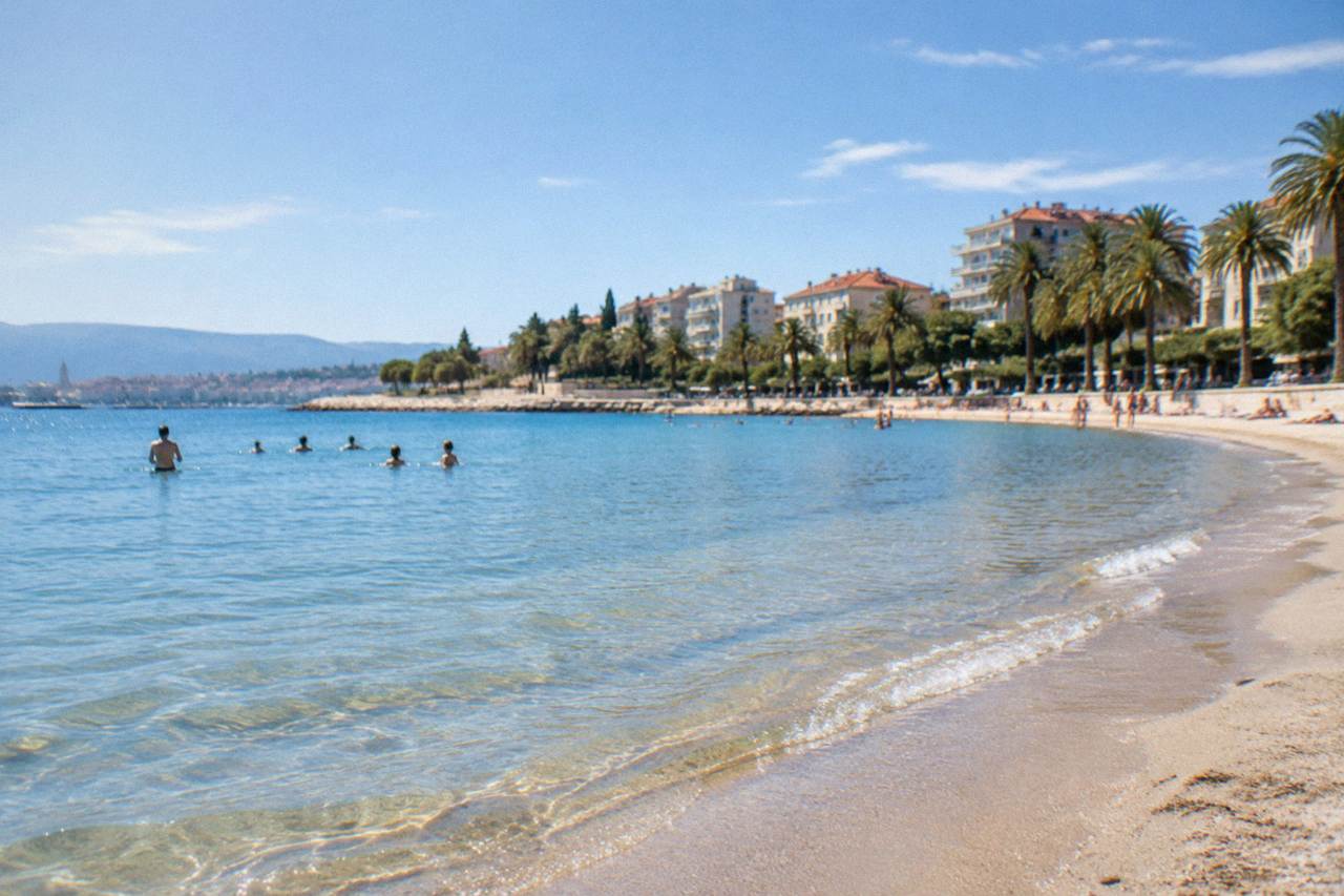 Sandy beach with shallow water and palm trees beside apartment buildings on a sunny day
