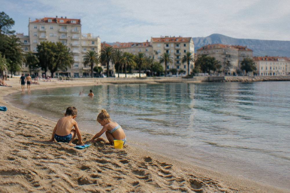Children playing on sand beside shallow water with apartment buildings behind