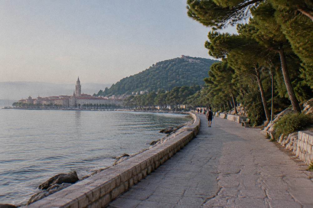 A wide coastal walkway leading from the old town waterfront towards the wooded Marjan hill