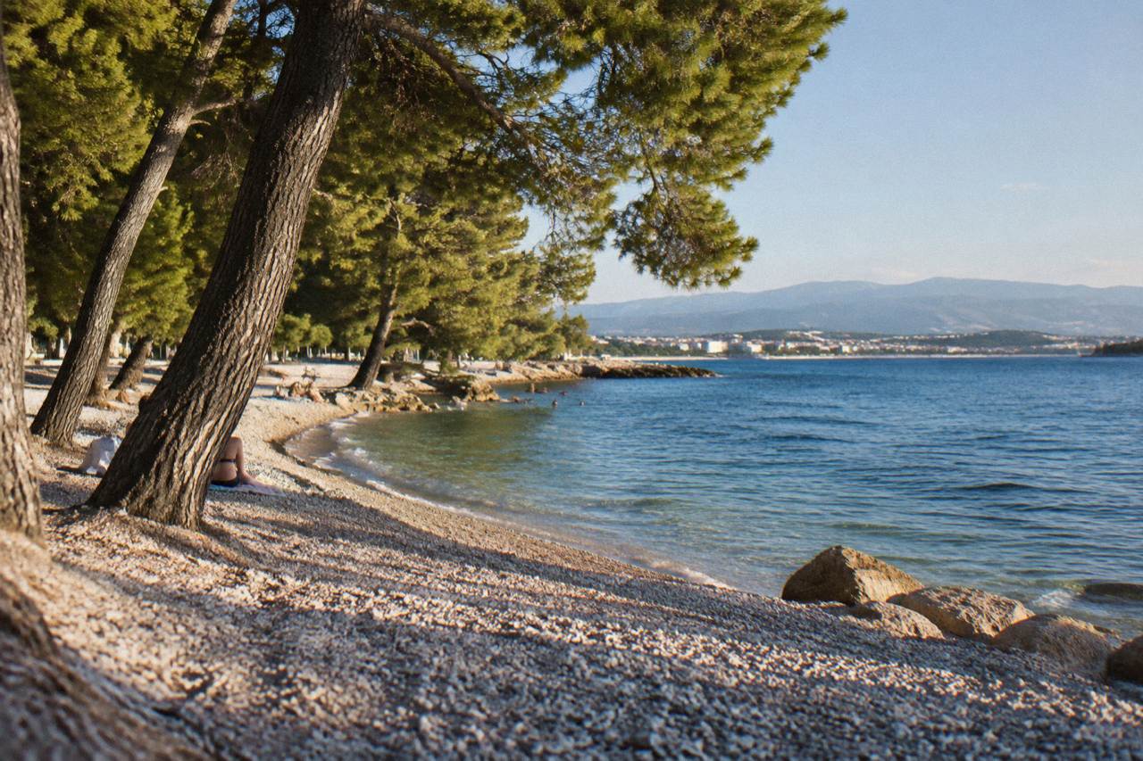 A calm pebble beach with pine trees reaching the waterline in the Stobrec district of Split