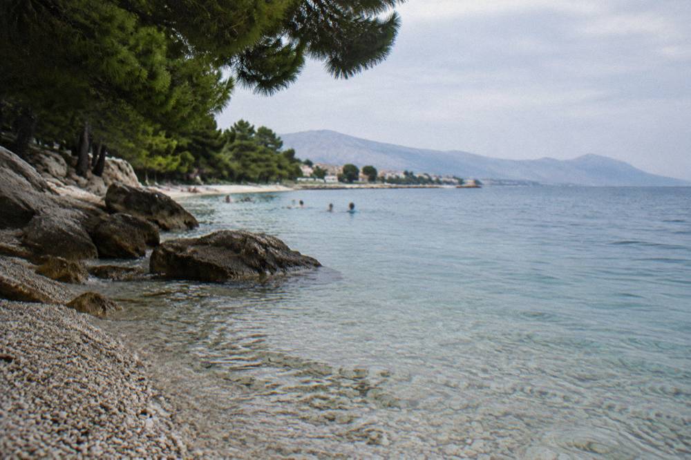 Clear shallow water over pebbles beside a pine shaded swimming spot in Stobrec
