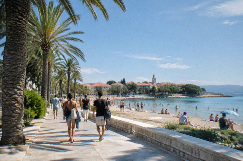 Palm-lined seafront promenade with people walking toward a sandy beach