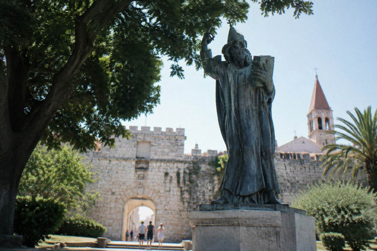 A large bronze statue of a robed medieval bishop standing on a pedestal beside an old stone city gate in Split