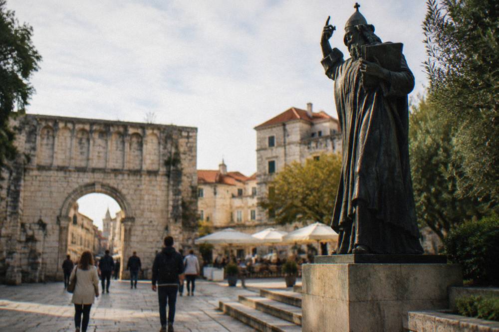 People walking past the Golden Gate stone arch of Diocletian's Palace toward a small square with a cafe terraces