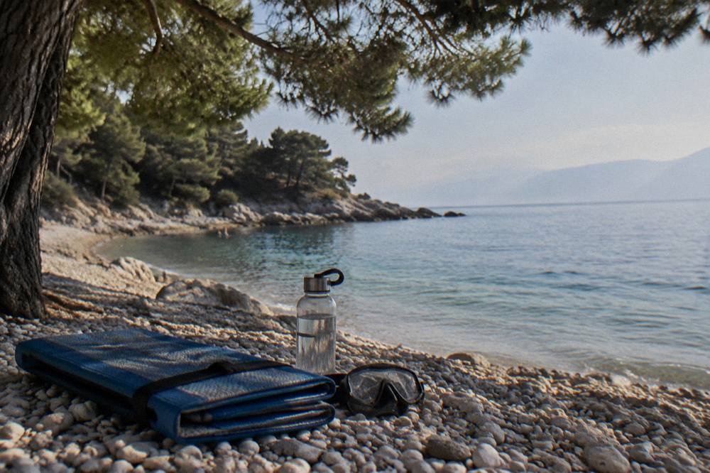 A folded beach mat, mask and water bottle set on smooth pebbles under a pine tree