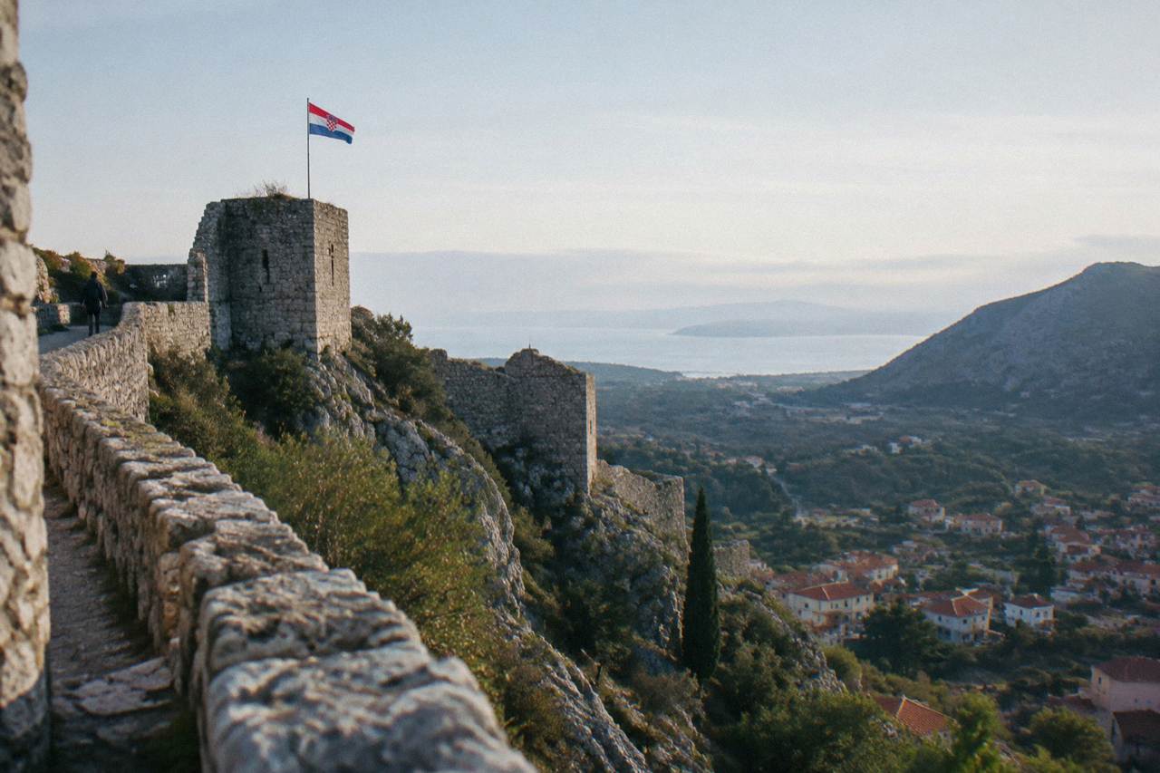 A stone fortress wall on a rocky ridge above a village and hills near Split