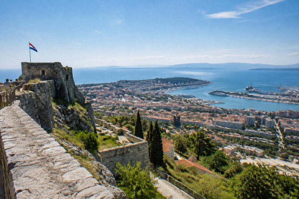 A wide view from fortress walls across a coastal city, its rooftops and the sea beyond