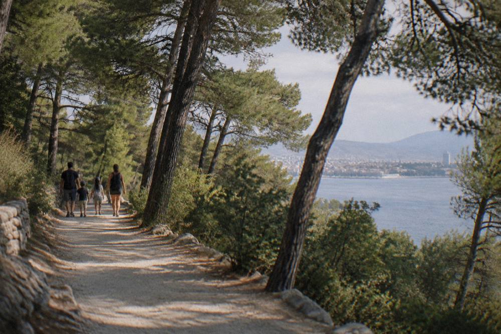 A family walking a shaded gravel path through pine forest on Marjan hill in Split