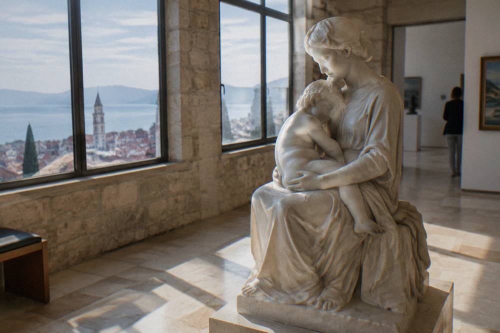 A marble sculpture of a seated mother and child in a sunlit gallery room