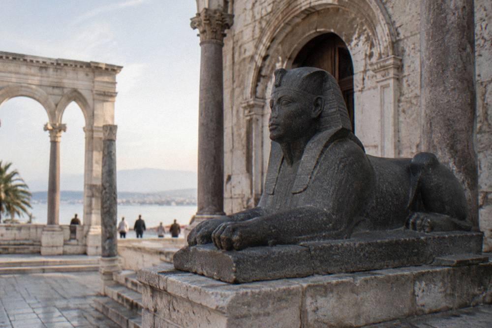 A dark granite Egyptian sphinx on a stone base beside an arched Roman doorway