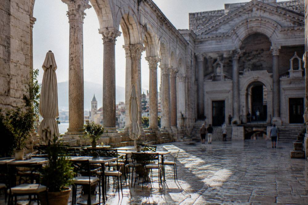 A stone-paved square with arched Roman columns and café tables in morning sunlight