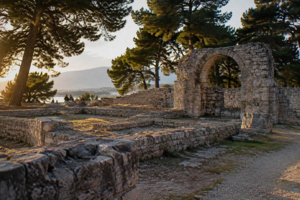 Low Roman wall foundations and an arched stone structure surrounded by pine trees at Salona