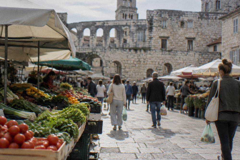 Shoppers carrying bags walking between produce stalls in front of an old stone palace wall