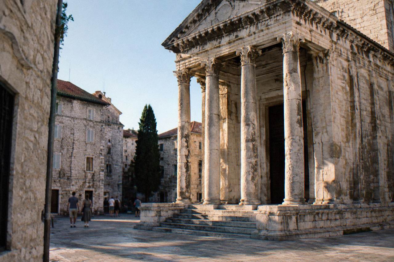 A weathered stone Roman temple facade with carved columns standing in a narrow square inside an old town