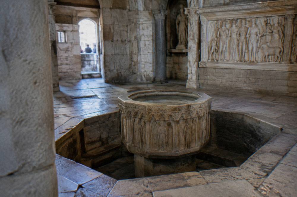 An interior of a small ancient stone building with a sunken baptismal font and carved reliefs