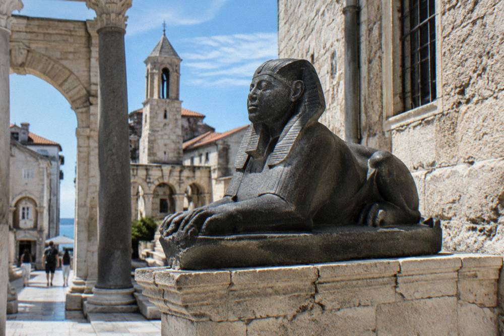 A dark granite sphinx statue on a plinth beside an old stone building
