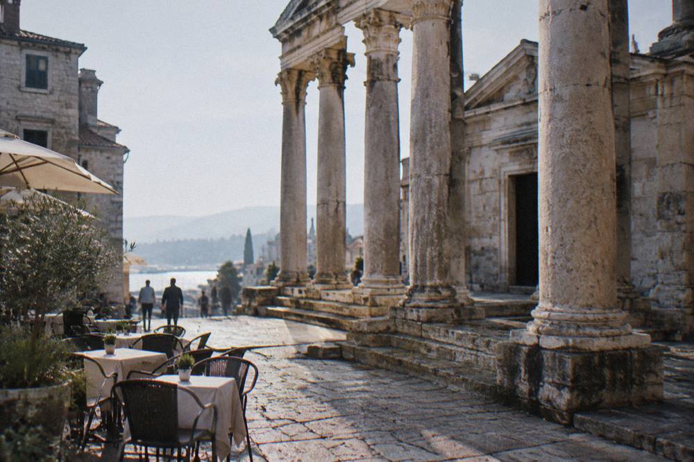 A sunlit ancient colonnaded square with stone columns and cafe tables in a Mediterranean old town