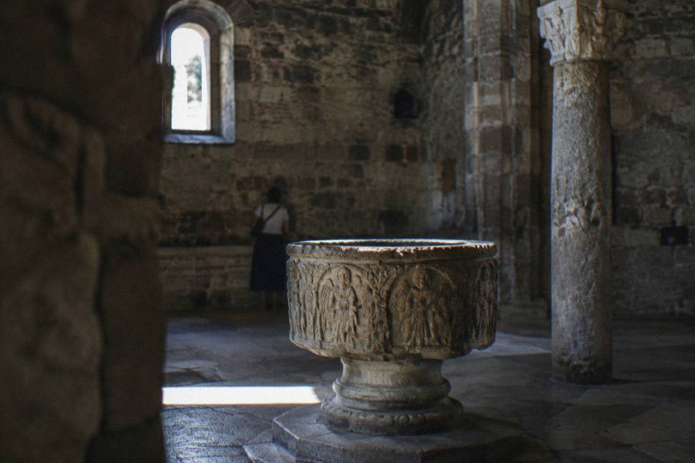 An ancient stone baptistery interior with a carved font lit softly through a small window