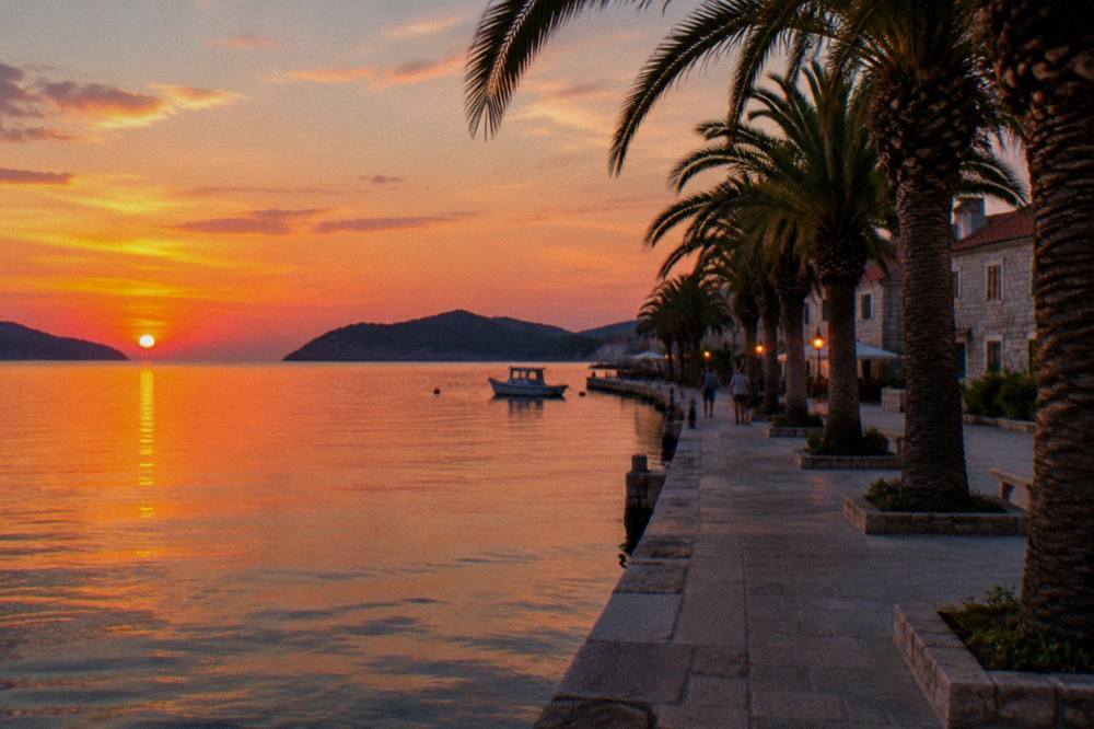 A palm-lined seaside promenade at dusk with stone buildings behind