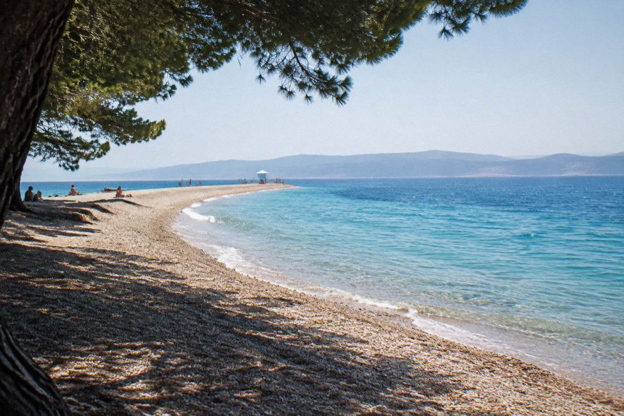 A long pebble cape reaching into clear blue Adriatic water under a sunny sky