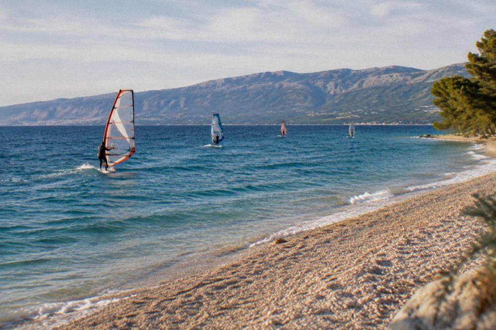 Windsurfers on blue water near a pebble shoreline in afternoon light