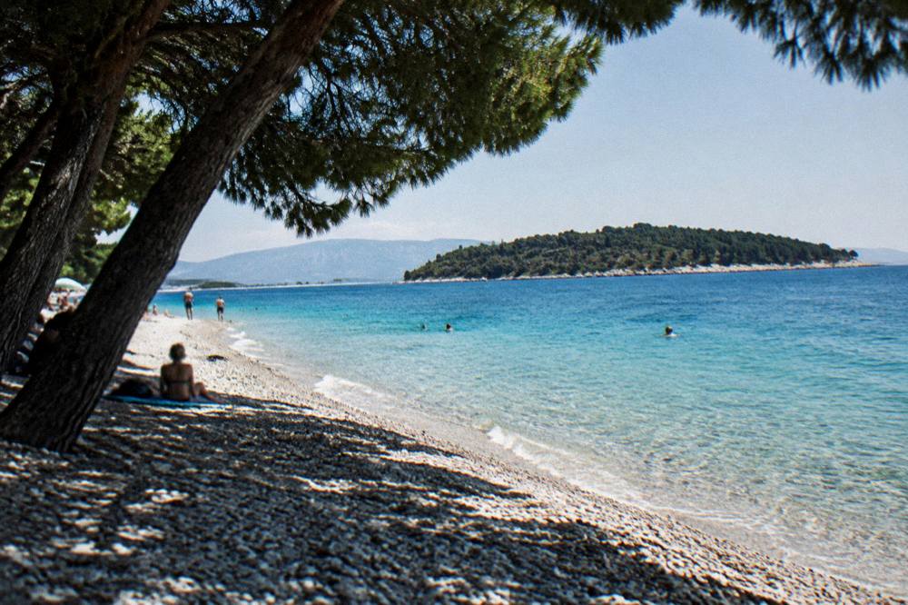 A pebble beach reaching into turquoise shallows with a wooded island in the distance