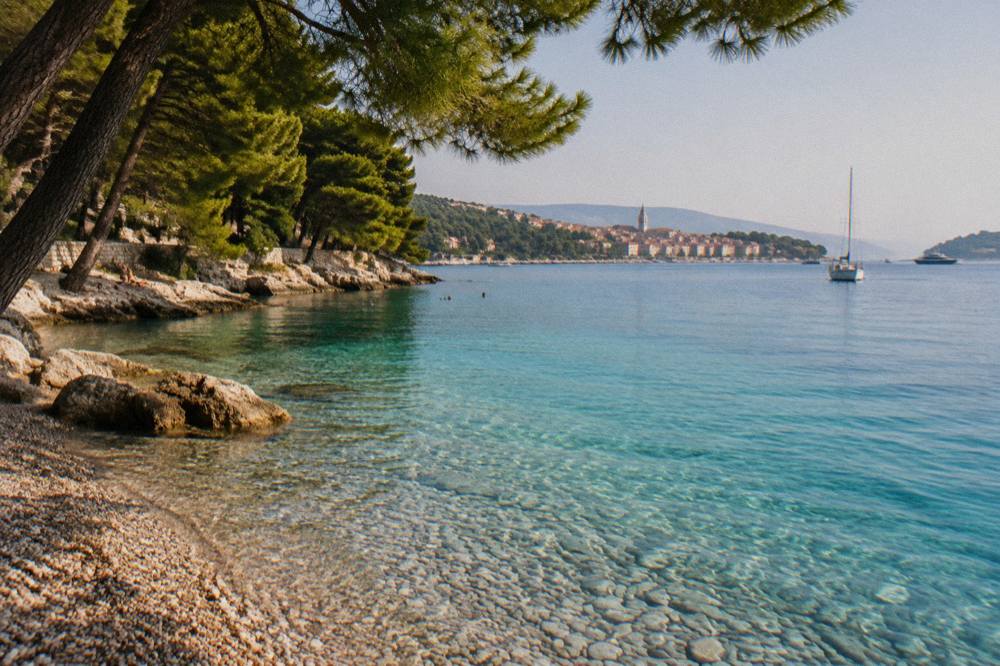 Clear turquoise water over a pebble beach with pine trees reaching to the shoreline