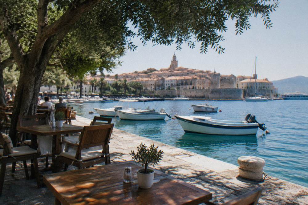 Outdoor cafe tables beside a stone quay with moored boats and clear water beyond