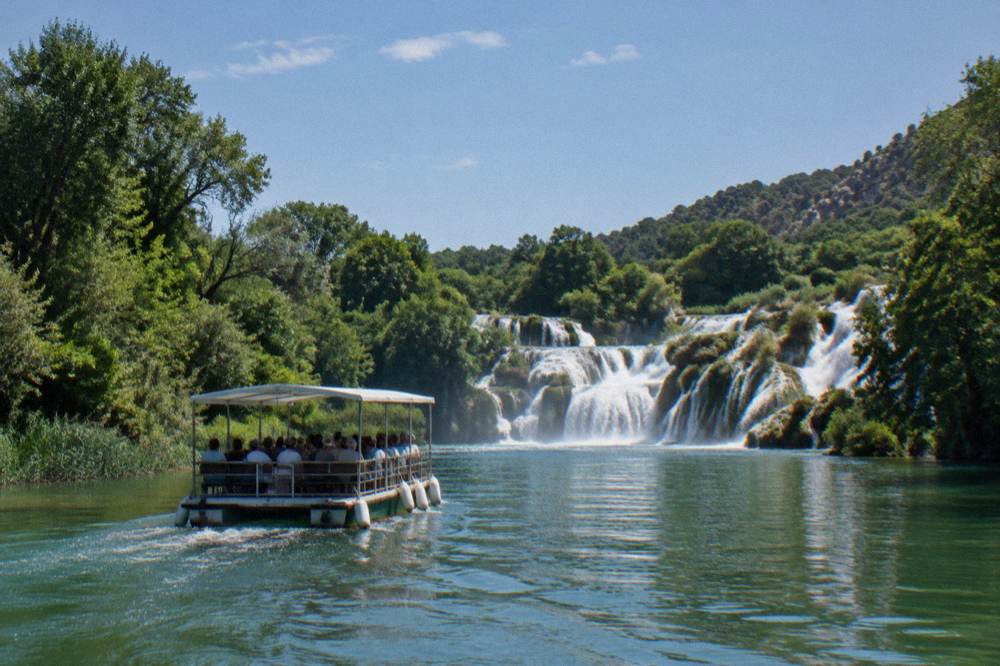 Boat carrying visitors along a calm green river toward waterfalls