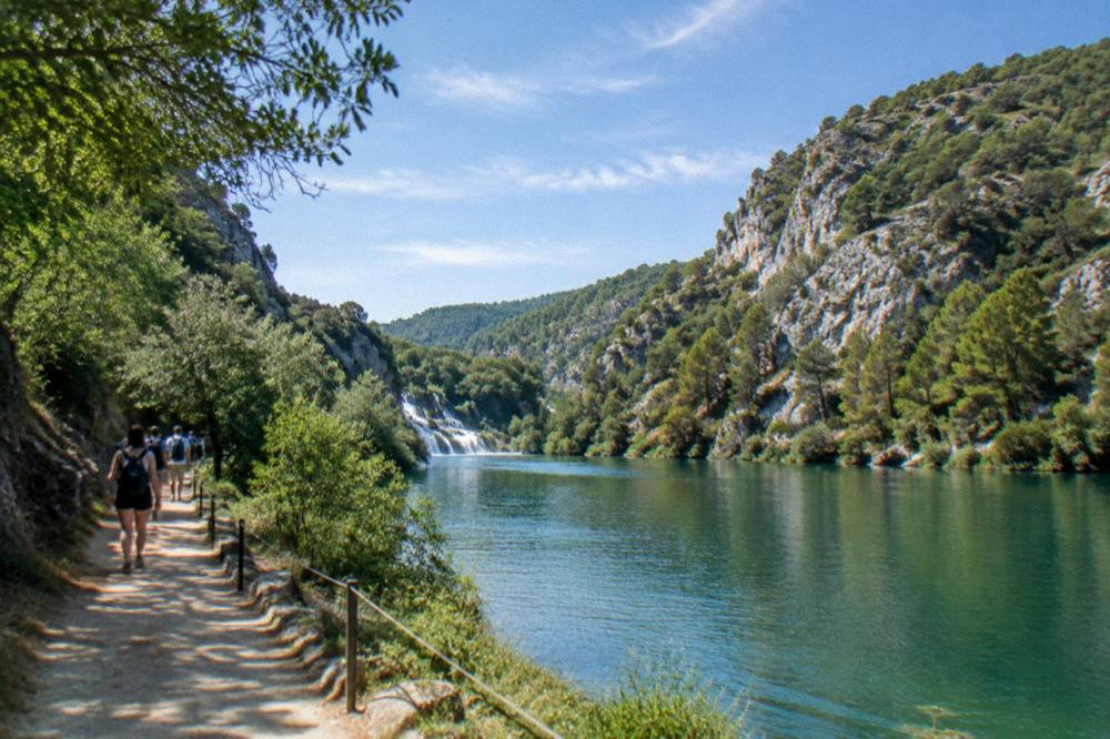 Visitors walking a riverside trail beside calm green water in a canyon