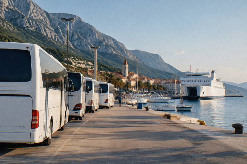 A row of white buses parked at a seaside bus station next to a ferry port with moored ships