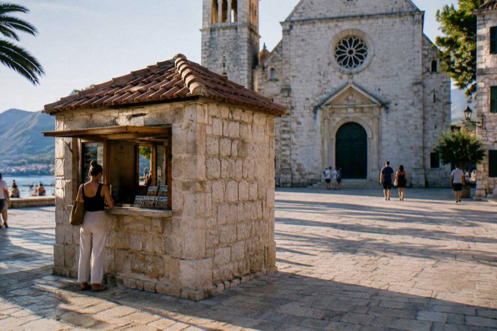 A stone kiosk beside a sunlit square selling tickets to visitors outside an old church facade