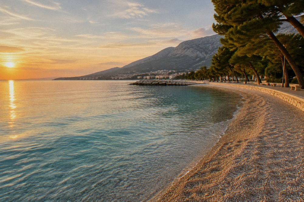 An empty pebble beach at golden hour with calm turquoise water and a tree-lined promenade behind it