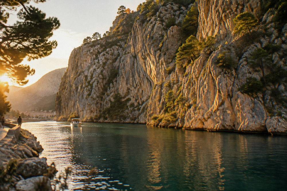 A tall limestone gorge wall rising above calm green river water with pine bushes on ledges