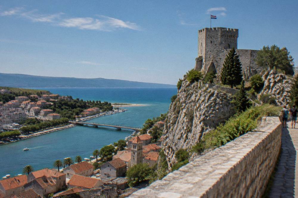 A stone fortress tower on a rocky outcrop above red rooftops and a river mouth