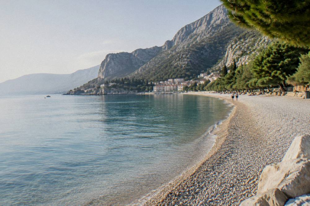 A pebble beach curving along a coastline with wooded mountains rising behind it