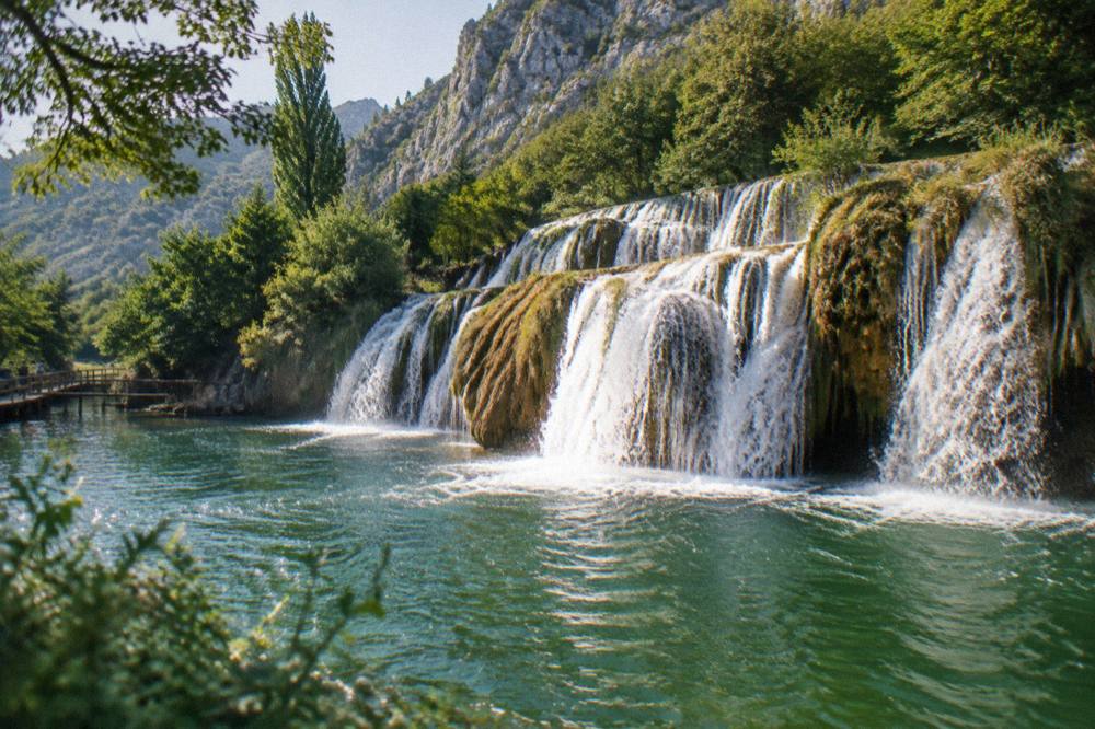 A wide waterfall spilling over travertine barriers into a green lake in a forested park