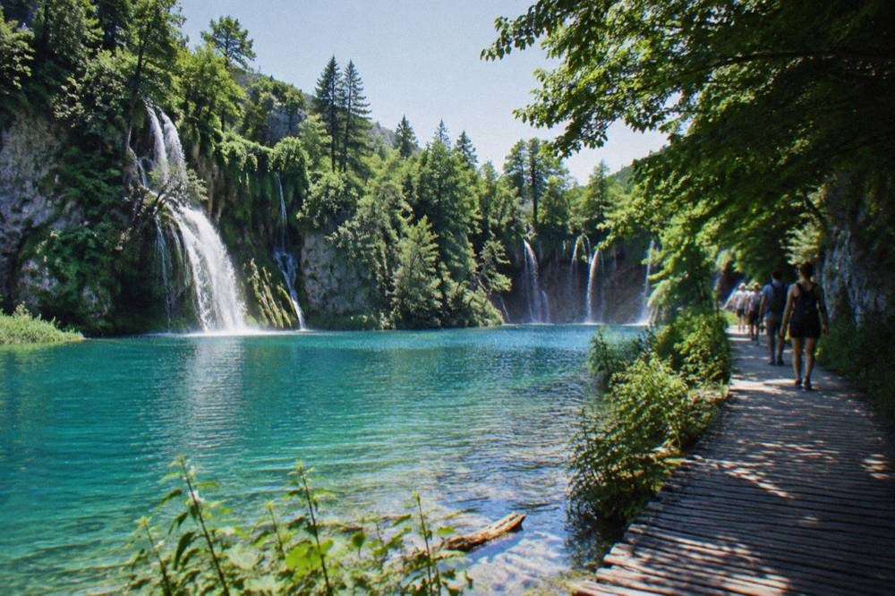 Visitors walking a wooden lakeside trail between waterfalls at Plitvice