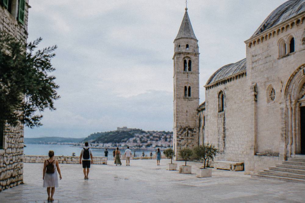 Travellers in light clothing walking a sunlit stone square beside a church tower