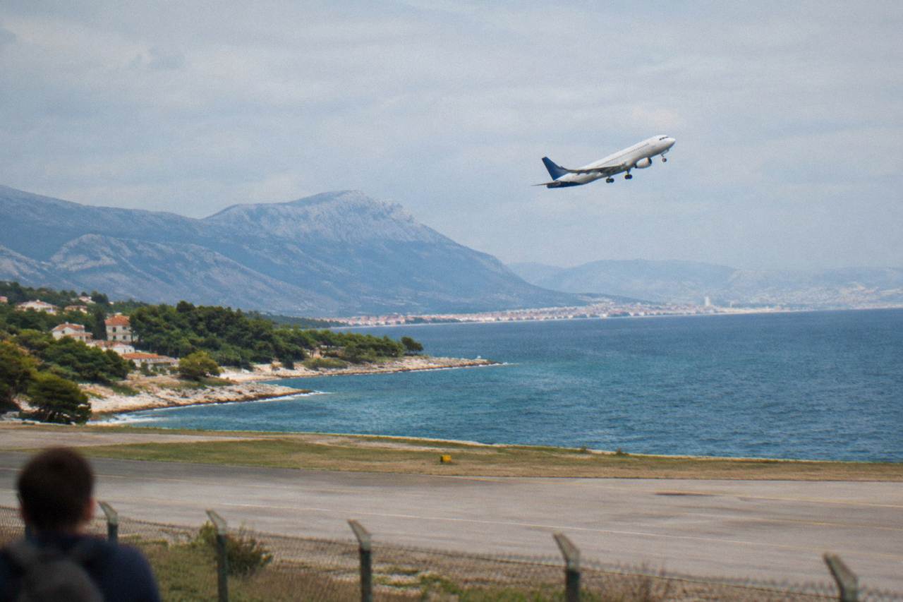 An airliner climbing over the Adriatic coastline near Split with rocky beaches and blue sea below