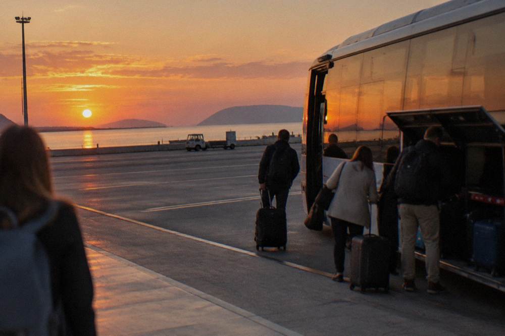 A shuttle bus parked at an airport kerb with passengers loading luggage in warm evening light